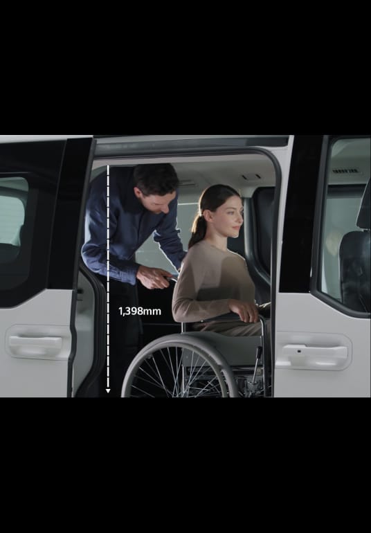 Ein Rollstuhlfahrer steigt mit Unterstützung einer weiteren Person im Fahrzeuginneren durch die Seitentür in den Kia PV5 WAV ein.